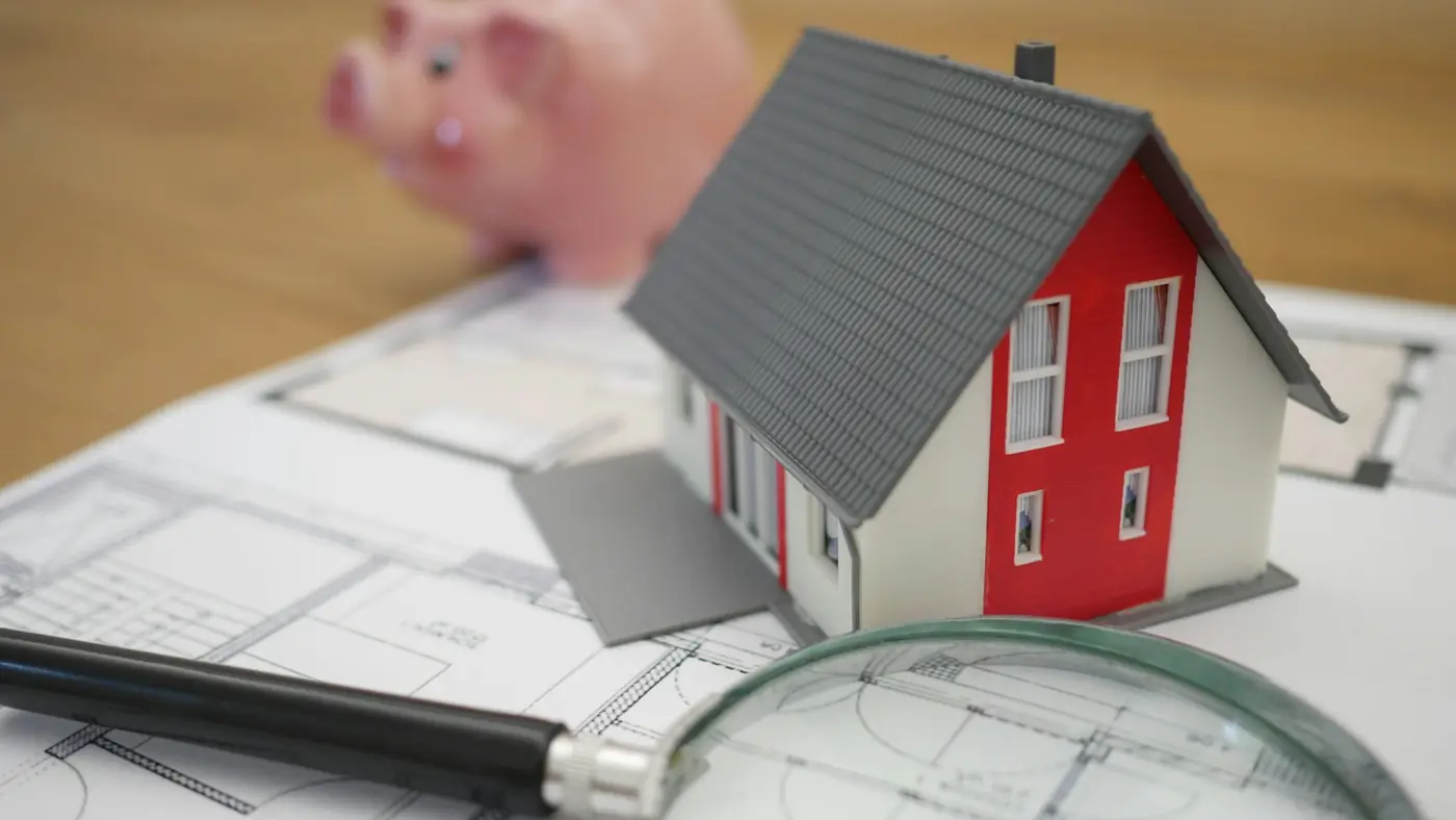 A model house on top of house plans, with a magnifying glass and a piggy bank