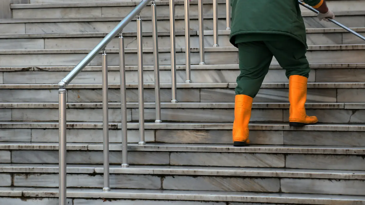 Un trabajador con ropa verde y botas de goma anaranjadas en unos escalones de piedra mojados, junto a una baranda de metal